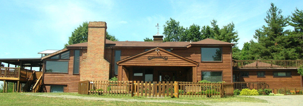 wooden fence and wooden building with large windows and 2 wooden decks with large evergreen trees behind. Chimney in front.