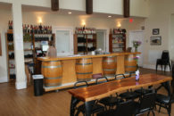 Room with table and bar with lady standing by shelves full of bottled wines.