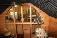 Round window looking into room with stainless wine making equipment, oak barrels on rack