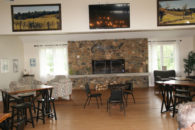 Large room with wood floor and stone fireplace with Casanel sign over top in between windows with sheers