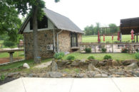 Small stone building with metal roof beside patio with tables an red umbrellas