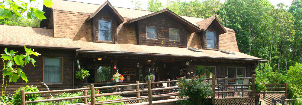 Wooden 2-story house with ramp and railing surrounded by woods with tall trees, garden next to ramp.