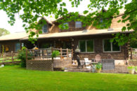 Front view of 2-story wooden house with ramp, pation with furniture, Furniture sitting on mulch by fire pit.