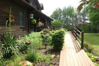 Front view of wooden ramp with railings leading to wooden house with garden on left and woods in the distance