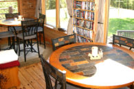 Brightl room with 2 round high tables with black charis, view of patio and yard, shelf full of books, wooden floor.