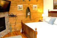 Dark oak queen bed with white coverings, wooden wall with pictures, wooden floor with rug, glat tv and gas fireplace in wall