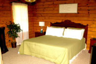 Queen bed with scalloped header with gold bed covering, wooden walls, window with sheers, tan carpet, nightstands.